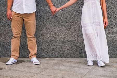 Man in white shirt and woman in white dress holding hands of each other while standing near the grey wall.の写真素材