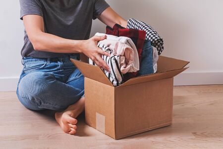 Unrecognizable woman putting clothes in cardboard box while sitting on the floor. Donation or packing for moving stuff in new place.の写真素材