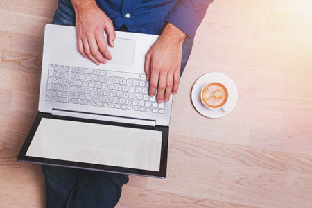 Man sitting on the wooden floor and working on laptop with a cup of coffee near him, view from the...
