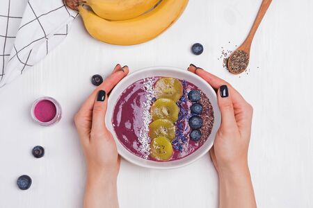 Woman's hands holding acai berry smoothie bowl topped with seeds, flowers and blackberries on white table, top viewの写真素材