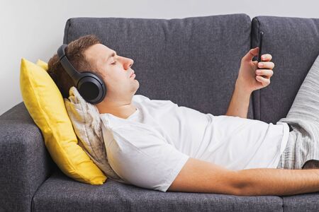 Man in wirwlwss headphones watching lying on sofa and looking on his smartphoneの写真素材