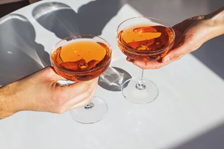 Close up view of man and woman hands holding glasses with cocktail of orange color on the white table in natural sunlight with shadowの写真素材