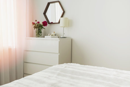 A part of modern bedroom in white colors with bouquet of peonies on the chest of drawers