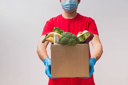 Delivery man wearing mask and gloves holding box full of different groceries.の写真素材