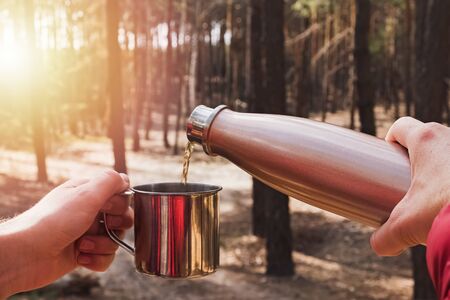 Man pouring tea from flask in a metal cup while camping in pine forest.の写真素材