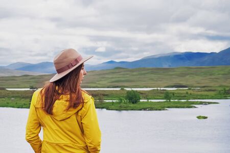 Young woman traveller in hat and raincoat standing near beautiful lake and looking in the distance, view from the back, travel concept.の写真素材