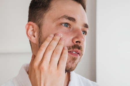 Handsome young man applying cream lotion on his face. Modern beauty and healthcare routine at homeの写真素材