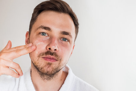Handsome young man applying cream lotion on his face. Modern beauty and healthcare routine at home