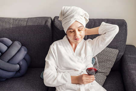 Young woman after shower in bathrobe with glass of red wine sitting in the living room.の写真素材