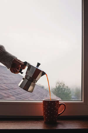Woman pouring coffee from moka pot in the cup standing near the windowの写真素材