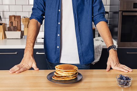 Man standing near the wooden table with a stack of freshly fried pancakes
