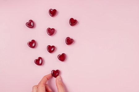 Hand holding one of scattered red jelly candies of heart shape on pink background