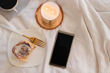Smartphone with empty screen on the bed with cinnamon bun and burning candleの写真素材