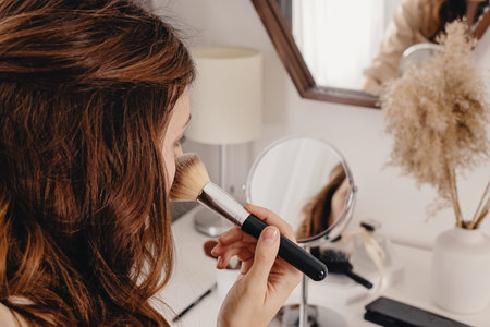 Young woman applying make-up to her face, looking at the round mirror