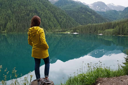 Woman traveler in yellow raincoat standing at the bank of peaceful mountain lakeの写真素材