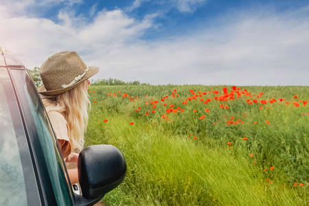 Young woman in a hat enjoying summer vacation. Looking from the window of a car to a green field with poppy flowers. Travel by car.の写真素材