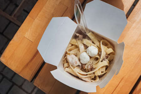 Tagliatelle pasta with mushrooms a paper box on wooden table of street cafe. Take away, delivery.の写真素材