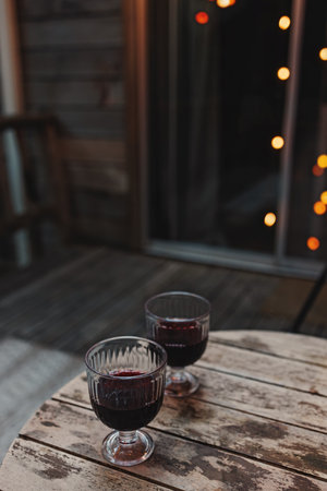 Two glasses of red wine standing on the wooden table with blurred trees and lights on the background. Summer cabin getawayの写真素材
