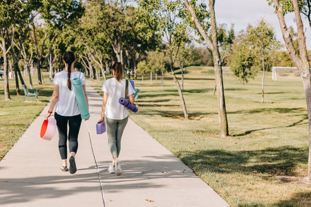 Two young women walking in the morning park, going to exercise outdoors. Carrying mats and other...
