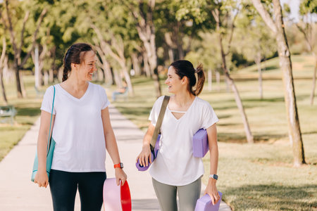 Two smiling young women walking in the morning park, going to exercise outdoors. Carrying mats and other equipment for yoga.の写真素材