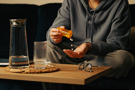 Unrecognizable man pouring tablets from orange plastic bottle into his hand, sitting on the couch at nightの写真素材