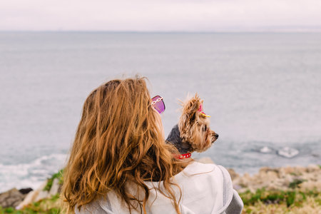 Woman holding a cute yorkshire terrier dog looking in the distance with ocean on the background, summer travel with dogの写真素材