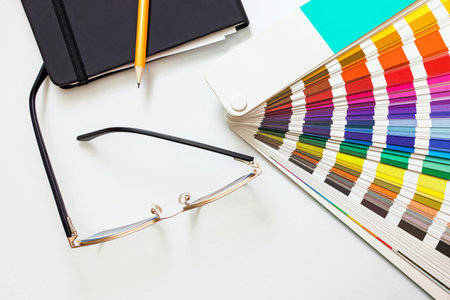 Graphic designer workplace concept, paint swatches color guide, sketchbook and glasses on white table, top viewの写真素材