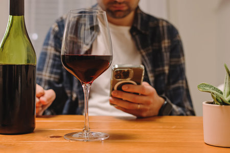 Unrecognizable man holding a phone and a glass of wine, using alcohol tracker app, mindful drinking.の写真素材