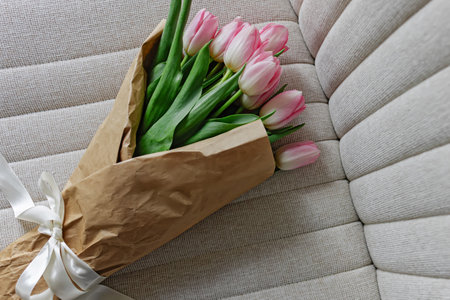 Bouquet of fresh pastel pink tulips wrapped in the craft paper decorated with satin ribbon lying on the beige chair,の写真素材