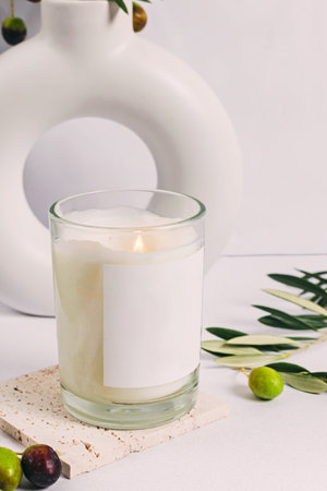 Burning aroma candle with empty white label on the table with olive branches and donut shaped vaseの写真素材