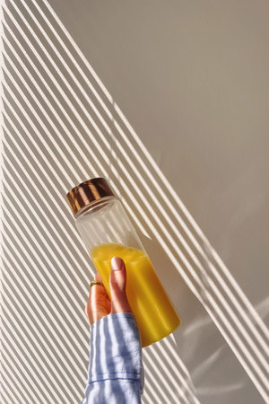 Female hand holding a reusable bottle with orange juice over the wall in the morning light and...