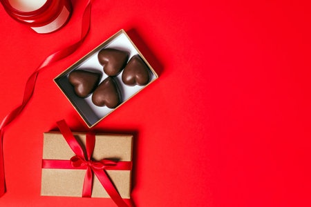 A gift box containing heart-shaped chocolates decorated with a red ribbon on a vibrant red background with a candleの写真素材