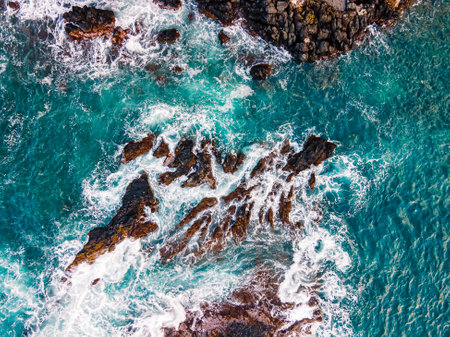 Aerial view of Turquoise waves crash against rocky coastline during sunny daylightの写真素材