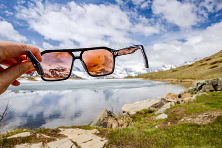 A person holds sunglasses in front of an alpine lake, showcasing the vibrant landscape through tinted lenses.の写真素材