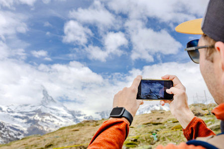 A person wearing an orange jacket takes a photo of a snow-capped mountain using a smartphone.の写真素材