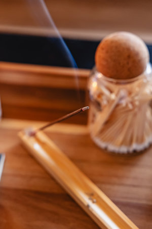 A close-up of a burning incense stick placed on a wooden holder, with gentle smoke rising against a blurred background, creating a calming atmosphere.の写真素材