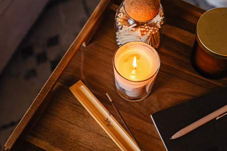 A warm and inviting scene featuring a lit candle on a wooden tray, surrounded by a jar of matches, a pen, and a notebook. The soft glow creates a relaxing atmosphere perfect for writing or reflection.の写真素材