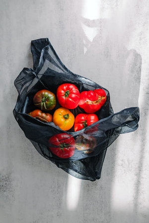 A variety of colorful tomatoes are displayed in a black mesh bag on a light textured surface. The vibrant reds, yellows, and greens of the tomatoes contrast with the dark bag, creating a visually appealing composition.の写真素材