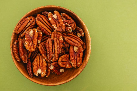 A wooden bowl filled with shelled pecans sits against a green backdrop.