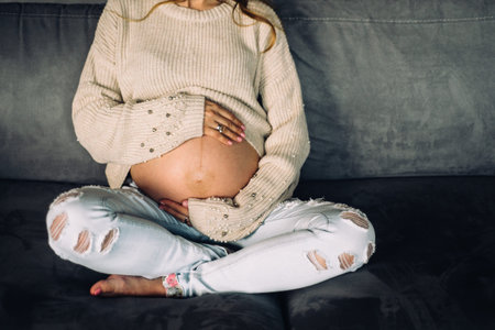 pregnant young woman is sitting on the sofa hugging belly with hands in jeans anf pullover