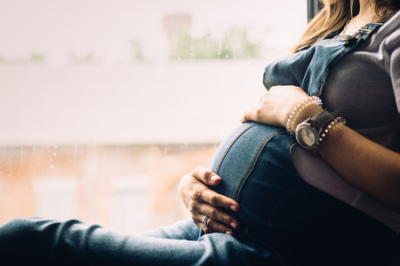 pregnant woman sitting on the window sill hugging belly with handsの写真素材