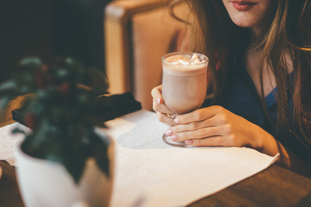 Beautiful brunette girl holding coffee liquid, sitting in front of unfocused plantの写真素材