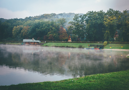 Mysterious lake and fall trees in hazy atmosphere, some buildings here and there also covered in grey fogの写真素材