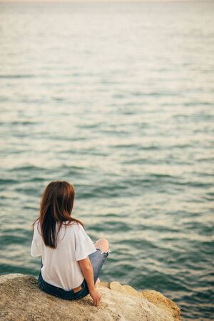 Beautiful young woman sits on the beach, dreaming and looking at the sea. A look from the back, against the background of sea and ocean waves on the sunsetの写真素材