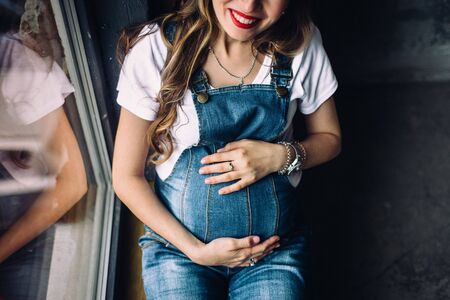 Smiling, pregnant woman, dressed in blue denim overalls, holding her hands on her tummy. She has beautiful smile and red lipstick on her lips.の写真素材