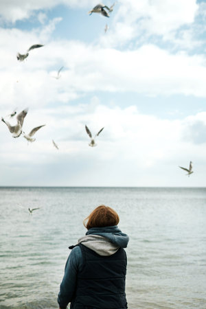 A lonely girl in a tank top and a pink sweater with a hood feeds seagulls by the sea.