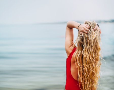  A young girl is standing in a red swimsuit near the sea. She hugs her head with hands and enjoys...