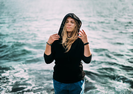A girl in a black sweater and blue jeans is standing by the sea with eyes closed and her arms are...