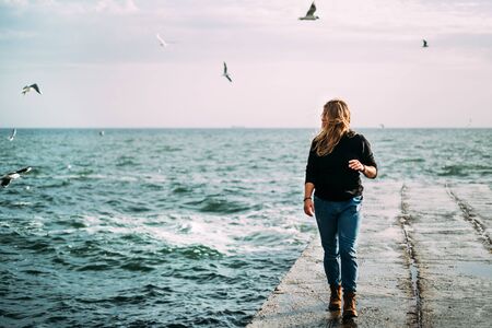 A girl walks on a pier by the sea with waves and a strong wind in a black sweater and jeans. Pink dawn and seagulls in the sky. Confidence in life. Determinationの写真素材