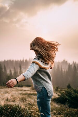 A girl stands on a mountain in a gray knitted sweater and blue jeans and looks into the distance. Wind blows hair. Dawn in the mountains. Pleasure. Peace and joy in travelの写真素材
